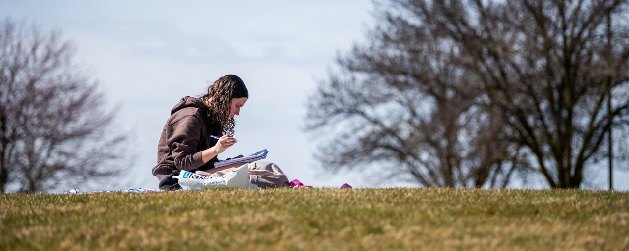 Students studies on a grassy hill with trees in the background